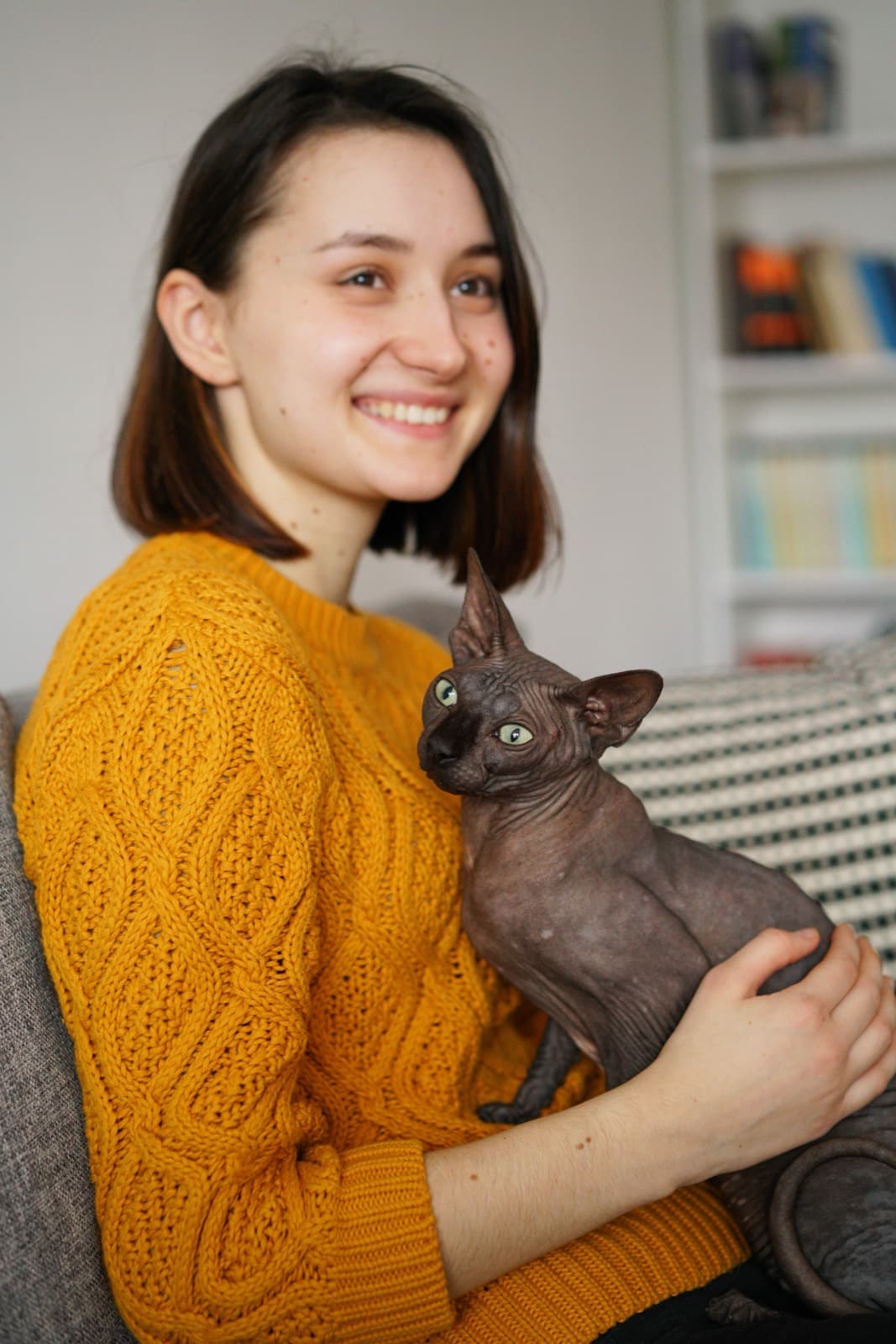 A smiling woman holding a dark grey Sphynx with green eyes on a sofa